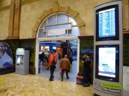 A NET ticket machine and a screen showing tram and bus departures in the railway station's ticketing area. The tram stop is through the arch and round to the left